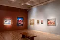 Six paintings hang in a gallery with one red wall and one white wall, with a bench in the center.