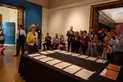 A group of people in a gallery look at a table with papers on it.