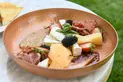 A low metal bowl filled with various sauces, cheeses, fruits, and cured meat.