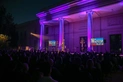 A crowd sits facing a large building with columns, lit in pir