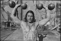 A grayscale image of a shirtless man, with long hair and a tattoo of a two-headed dragon, lifting barbells overhead.