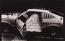 A grayscale image of a large, tiered wedding dress sticking out from the driver-side door of a sports car.