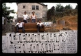 Four people sit on a couch on top of a wall, in a dry landscape, in front of a gray house; the house and wall are covered in graffiti, the largest tag reads 