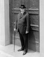 An older person dressed in a suit, holding a cane, stands in front of an intricatly-detailed door.