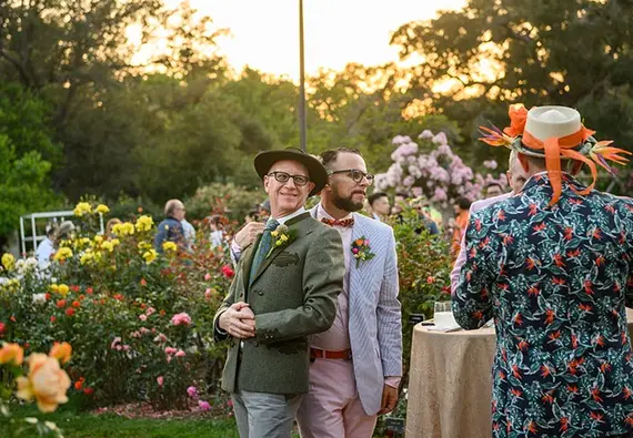 Two people in suits embrace in a rose garden.