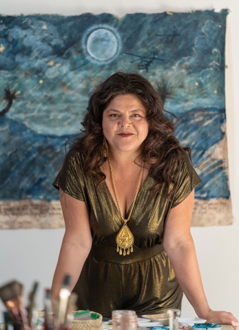 A woman stands behind paintbrushes in the foreground and a blue watercolor art piece behind her on the wall.