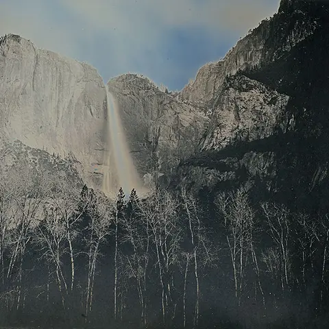 A waterfall cuts through a crevice in a tall mountain range, disappearing behind a stack of trees.