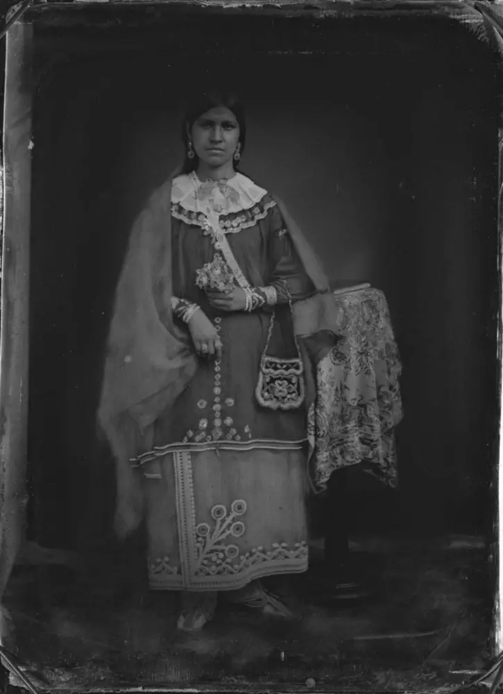 Black and white photograph of a Seneca woman. The woman wears clothing decorated in floral beadwork and she carries a bag decorated in floral beadwork.