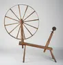 Large wooden spinning wheel with spokes attached to one end of a simple bench-like structure with a pulley device attached to the other end.  