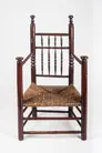 A large wooden armchair with original red stain, turned decoration on the chair back and arms, and a straw-colored, fibrous woven seat. 