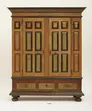 Large wooden wardrobe with panels, each with a border of red, green or black and painted decoration simulating the look of wood grain; drawers across bottom and cornice at top.