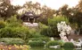 Garden with standing water in the foreground, a large rock next to the water ,and a one-room structure with a curved roof on a hill above the water.