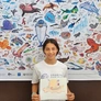 A young person holds a drawing on paper while standing in front of a poster filled with colorful illustrations of sea creatures.