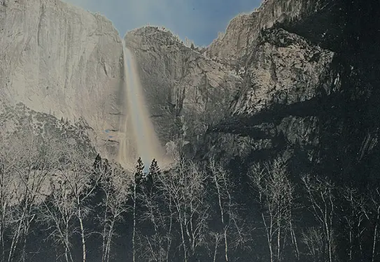 A waterfall cuts through a crevice in a tall mountain range, disappearing behind a forest of trees.
