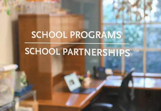 An open laptop is set on a desk seen through a glass window that says "School Programs" and "School Partnerships."