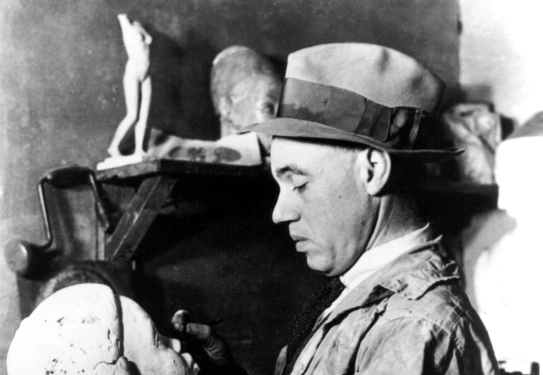 A light-skinned Black man wearing a painter's smock and a fedora stands and looks down at a sculpture of a head he is holding. The sculpture depicts a young Black person. Behind him, we see shelves with several sculptural pieces on them.