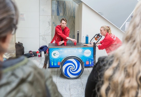 Two people in red coats running a science experiment in front of a crowd.