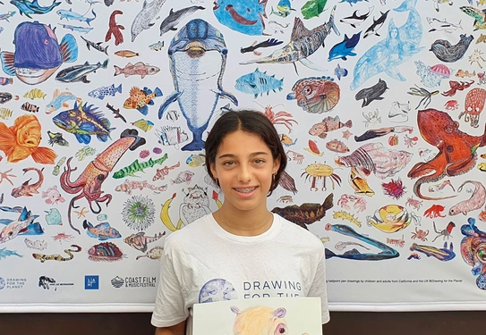 A young person holds a drawing on paper while standing in front of a poster filled with colorful illustrations of sea creatures.