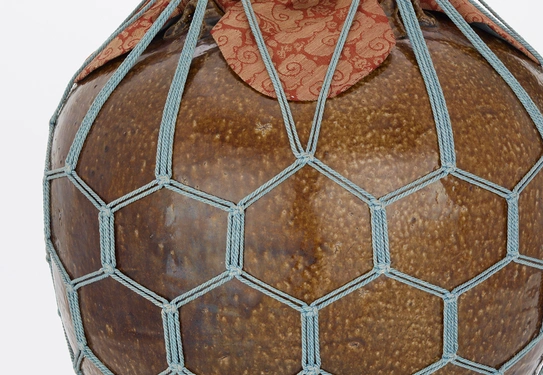 A brown ceramic jar with patterned cloth over the lid, the entire object is contained within a secure gray netting with a hexagonal pattern.