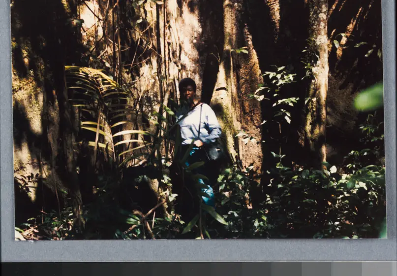 Octavia E. Butler stands in front of a large tree trunk. Green plants surround her.