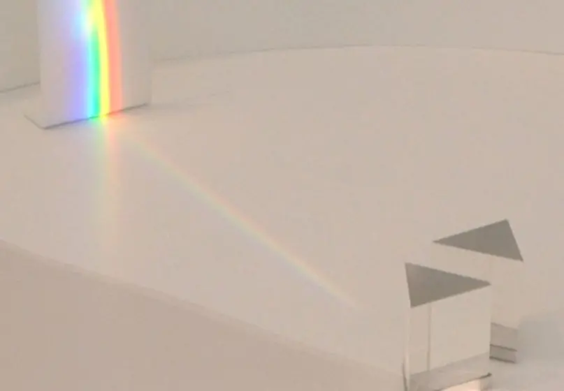 a beam of light shines through a triangle-shaped block of glass and lands on a white wall. The light displays as a rainbow on the white wall.