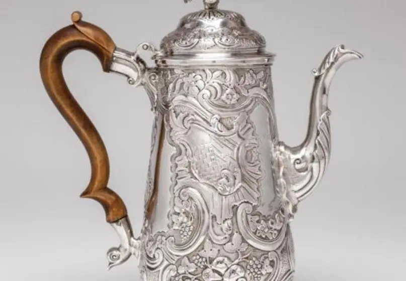 A decorated silver coffee pot with a wood handle.