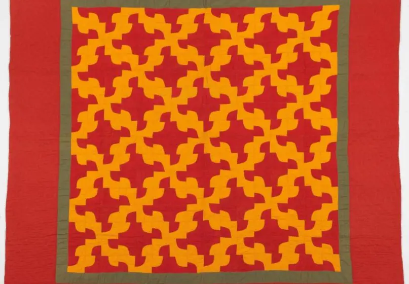 Red fabric quilt with square trimmed with green fabric at the center; the square contains a repetitive, symetrical pattern of a grid made of jagged red distorted diamond shapes, surrounded by curved pieces of yellow fabric.