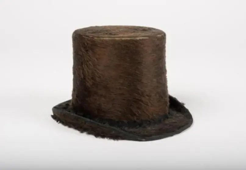 Miniature brownish-black top hat with furry texture, placed on a plain surface against a plain white backdrop. 