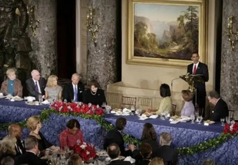 President Obama stands in front of a landscape painting as he gives a speech to a group of about twenty people.