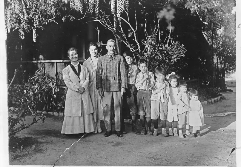 A black and white photograph of a middle aged man and woman stand with a young woman and six children. All of the people in the photograph are Japanese.