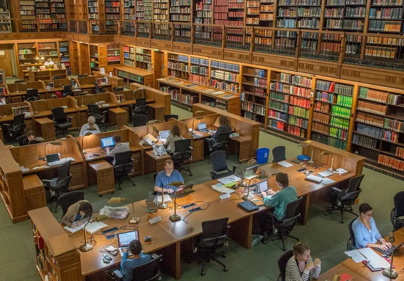 A large two story room with book-lined walls and long tables at which people work.