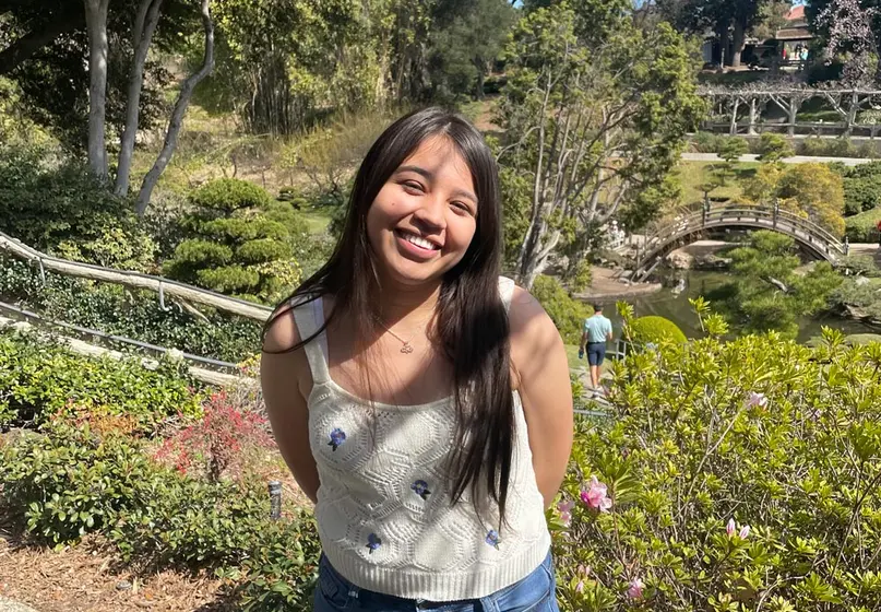 A college student smiles on a sunny day in a garden.