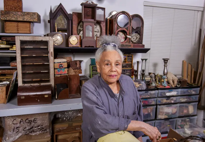 Color photograph of an older Black woman with white and grey hair pulled up to the top of her head in a short, curly ponytail. She is wearing a loose grey shirt and is standing in her artist studio, surrounded by wooden clocks, shelves, candlestick holders, and other miscellaneous items.