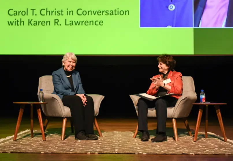 Two people sit in chairs on a stage. A large projection screen reads “Carol T. Christ in Conversation with Karen R. Lawrence.”