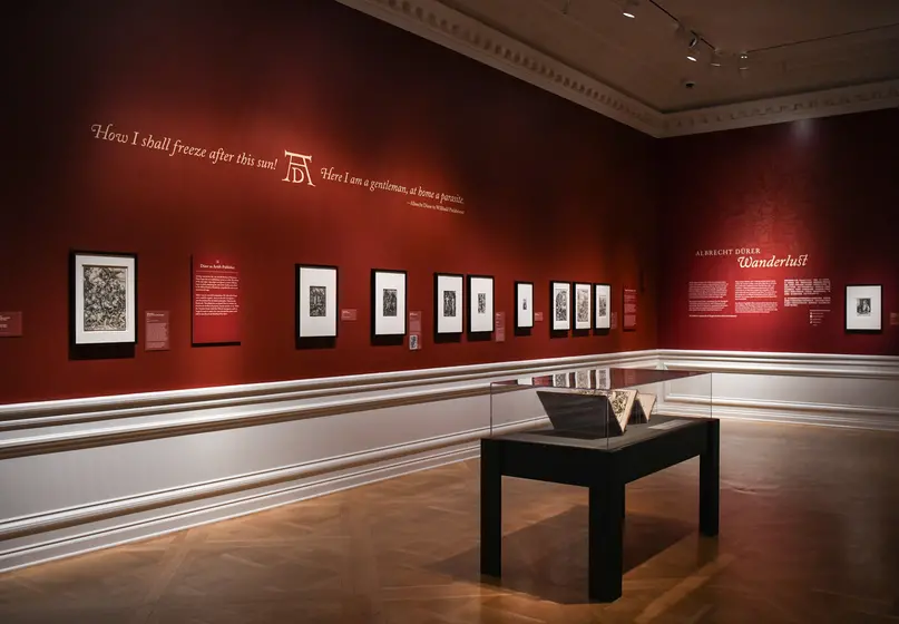 A gallery with dark red walls and beige text features matted artwork in black frames and a display case with two open books.