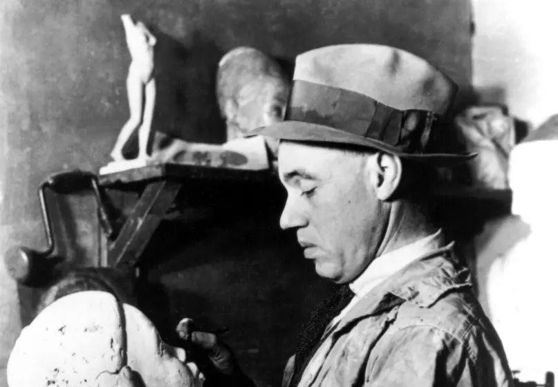 A light-skinned Black man wearing a painter's smock and a fedora stands and looks down at a sculpture of a head he is holding. The sculpture depicts a young Black person. Behind him, we see shelves with several sculptural pieces on them.