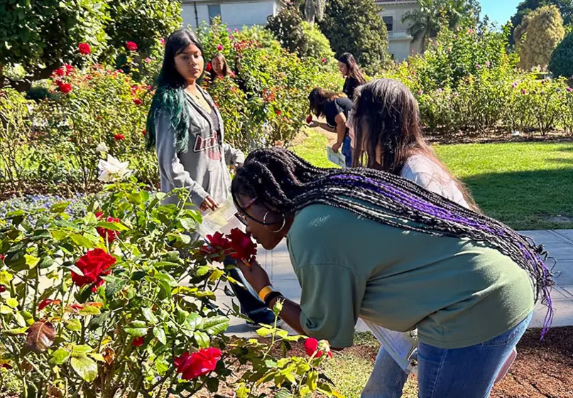 A young person leans in close to smell a red rose in a garden.