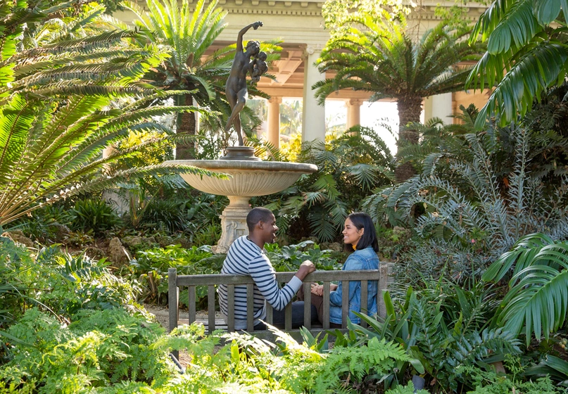 A couple sits on a bench near a fountain in a garden filled with tropical plants.