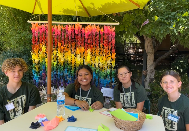 Four young people sit at a table with craft supplies, in front of a curtain of folded paper cranes.