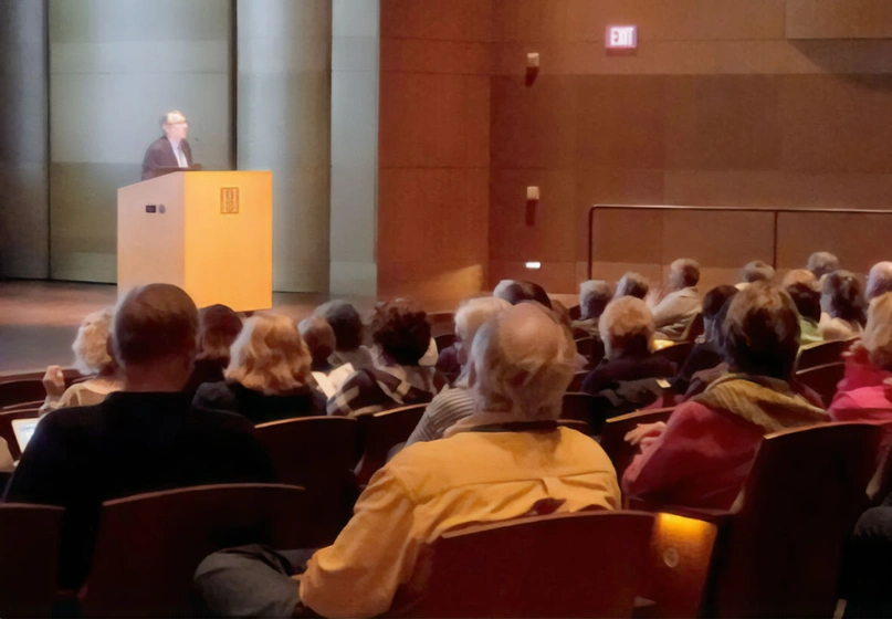 An auditorium audience watches a speaker at a lectern.