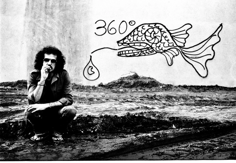 Grayscale image of a person squating on a dirt patch in front of a white wall with graffiti of a stylized fish and "360º."