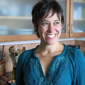 A person in a blue top smiles off camera while standing in a home kitchen.