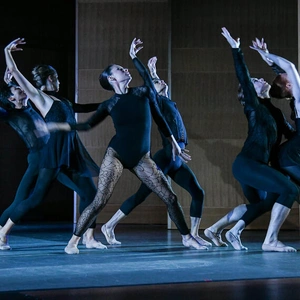 A group of dancers wear black outfits while performing on a dimly lit stage.