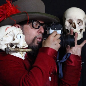 A person wearing a red blazer and a brown hat, holds a camera, with two skulls nearby.