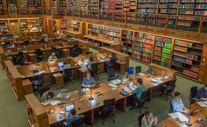 A large two story room with book-lined walls and long tables at which people work.