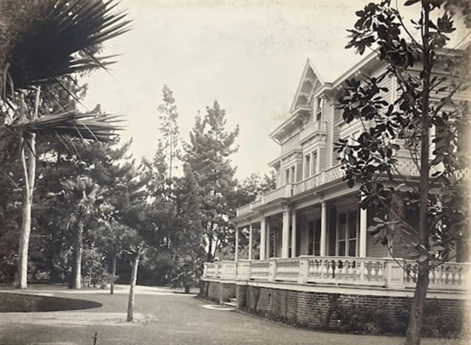 Black and white photograph of a large house with a wrapping porch. The house is surrounded by green grass and tall trees.