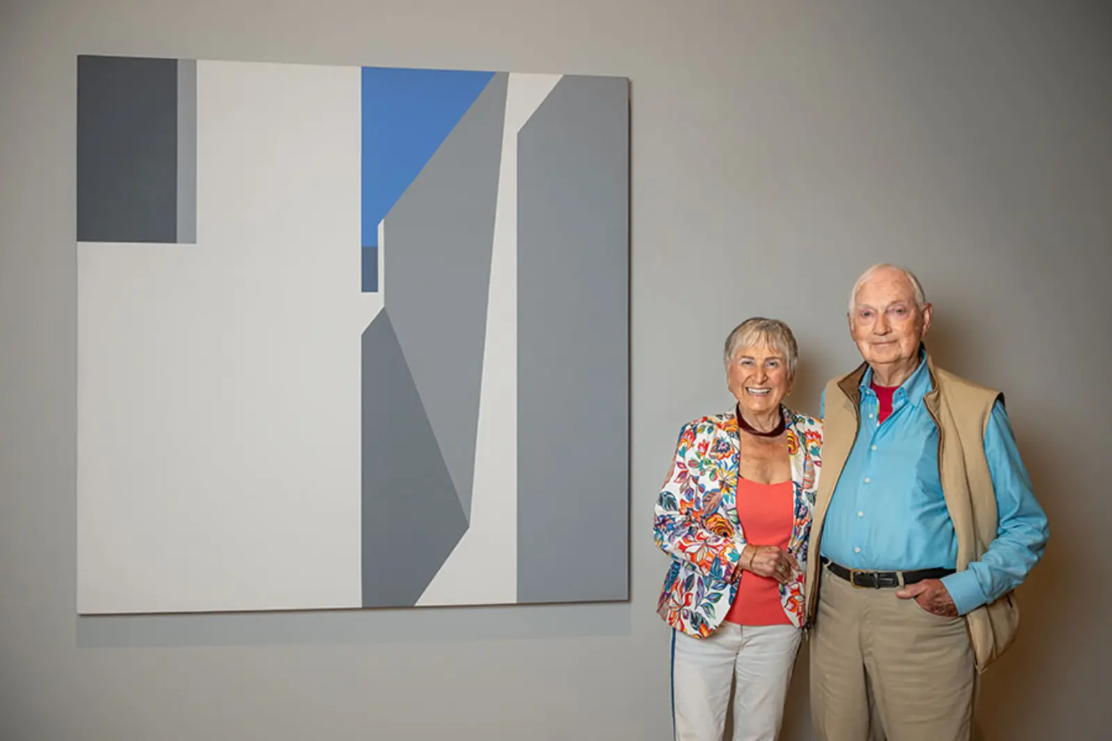 Two people pose next to an art piece on a wall.