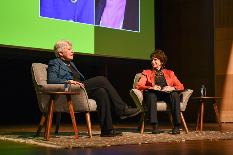 Two people sit in chairs on a stage, with a large projection screen in the background.