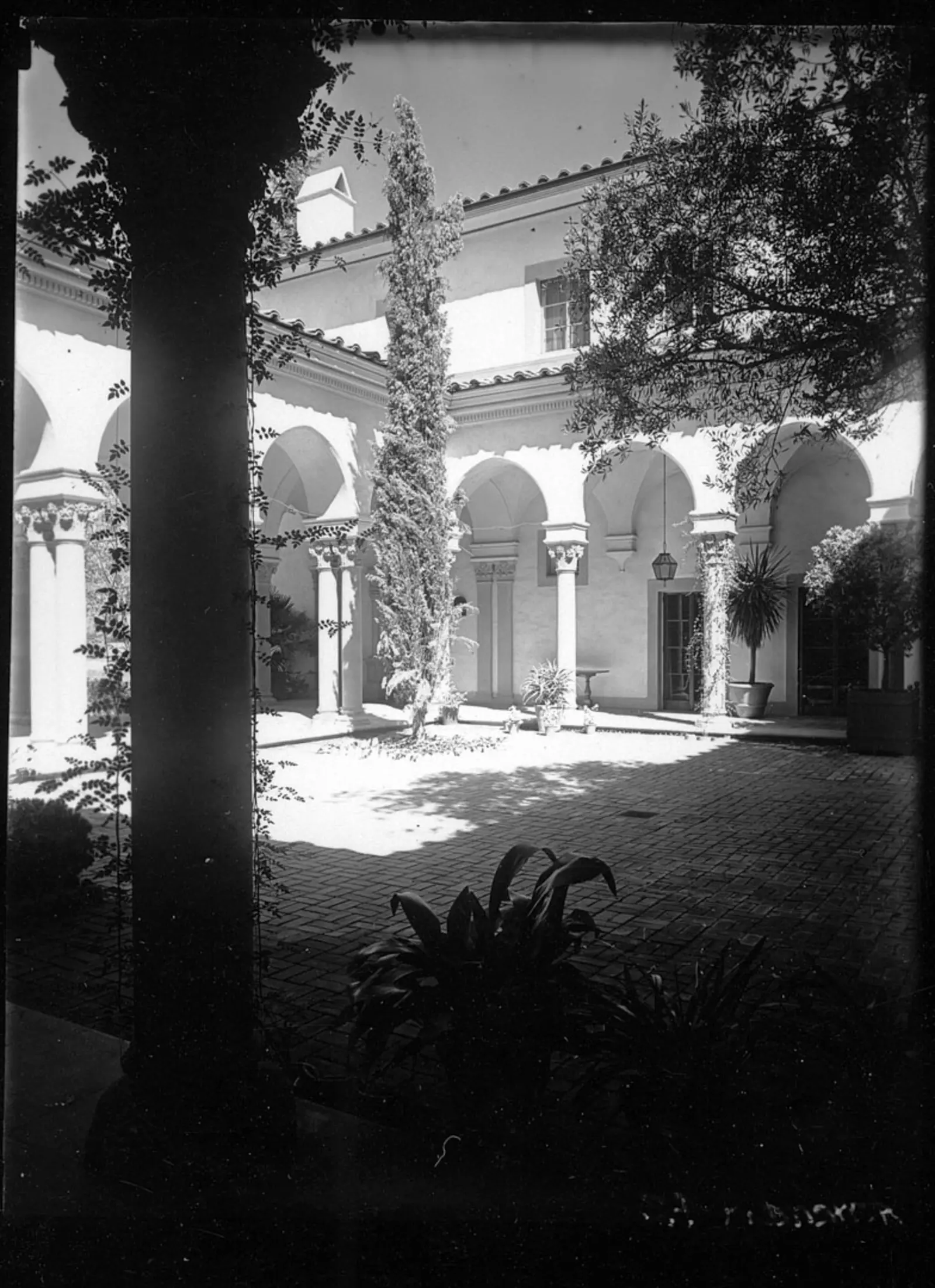 A black-and-white photo of an open courtyard with columns and arches, surrounded by foliage.