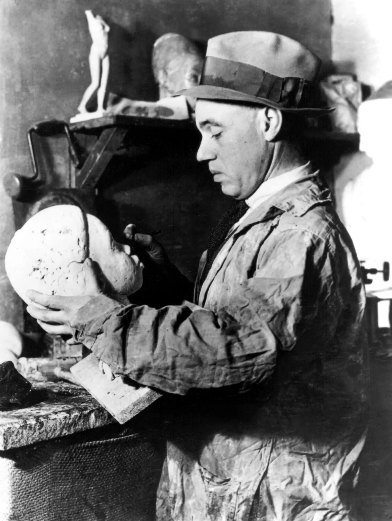 A light-skinned Black man wearing a painter's smock and a fedora stands and looks down at a sculpture of a head he is holding. The sculpture depicts a young Black person. Behind him, we see shelves with several sculptural pieces on them.